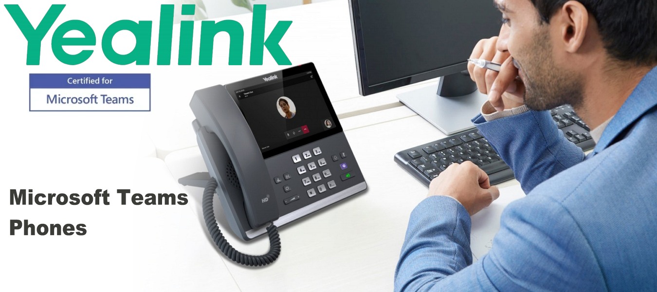 A man working at a desk with a Yealink phone displaying a Microsoft Teams call, keyboard, and computer monitor.